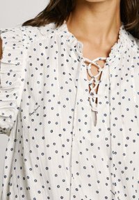 Blusa bianca con pois navy, maniche a balze, scollo con lacci e sottili strisce verticali lucide, indossata da una persona con capelli castani di lunghezza spalla.