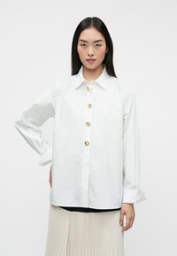Camisa blanca de botones con cuello, con botones circulares dorados y mangas largas, combinada con una falda clara con pliegues.