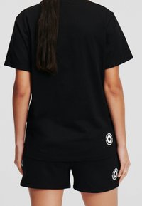 Camiseta de manga corta negra de algodón, con un dobladillo redondeado y un detalle de logo circular en la parte inferior izquierda. Combinada con pantalones cortos negros.