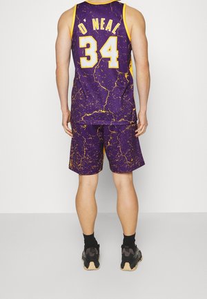 Mann, der ein lila-gelbes Basketballtrikot und Shorts mit der Nummer 34 und dem Namen O'Neal trägt, steht mit dem Rücken zum Betrachter vor einem einfarbigen Hintergrund.