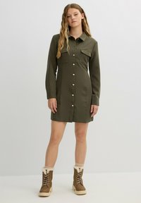 Vestido camiseros verde oliva con mangas largas, cierre de botones, dos bolsillos en el pecho y un corte entallado, combinado con botas marrones de cordones.