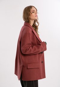 Przesadny bordowy wełniany żakiet z klapami, dwiema guzikami i jedną kieszenią na piersi. Charakteryzuje się gładką fakturą i zarysowanym kształtem.