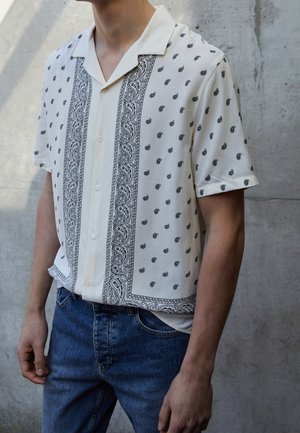 Mann trägt kurzärmeliges cremefarbenes Hemd mit schwarzem Paisley-Muster und blaue Jeans, steht vor einer grauen Betonwand.
