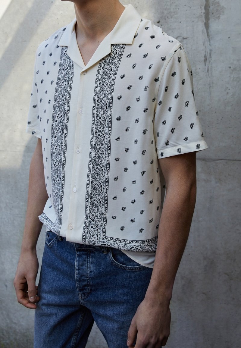 Homme portant une chemise crème à manches courtes avec un motif cachemire noir et un jean bleu, debout contre un mur en béton gris.