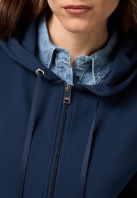 Marinblå zip-up hoodie med dragsko, lager över en ljusblå denimskjorta med spetskrage och tryckknappar.