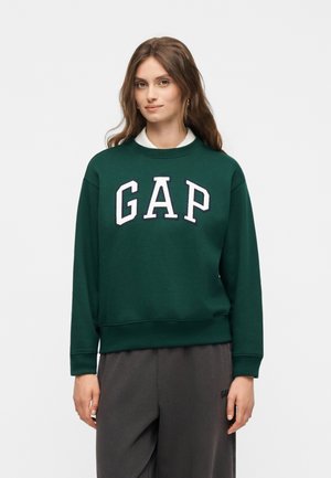 Fiatal nő sötétzöld GAP pulóverben és sötétszürke nadrágban, egyszerű világos háttér előtt áll.