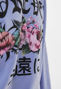 Sweat-shirt violet clair avec du texte noir et des motifs floraux rose et vert, mettant en valeur un tissu texturé avec des impressions graphiques détaillées.