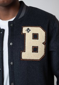 Person wearing a dark navy varsity jacket with a large beige letter B patch bordered in brown on the chest, over a white shirt.
