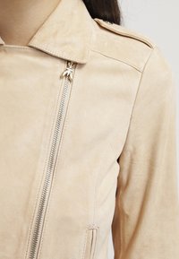 Giacca in suede beige con zip argentata asimmetrica, colletto, spalline e tasca verticale sul lato destro.