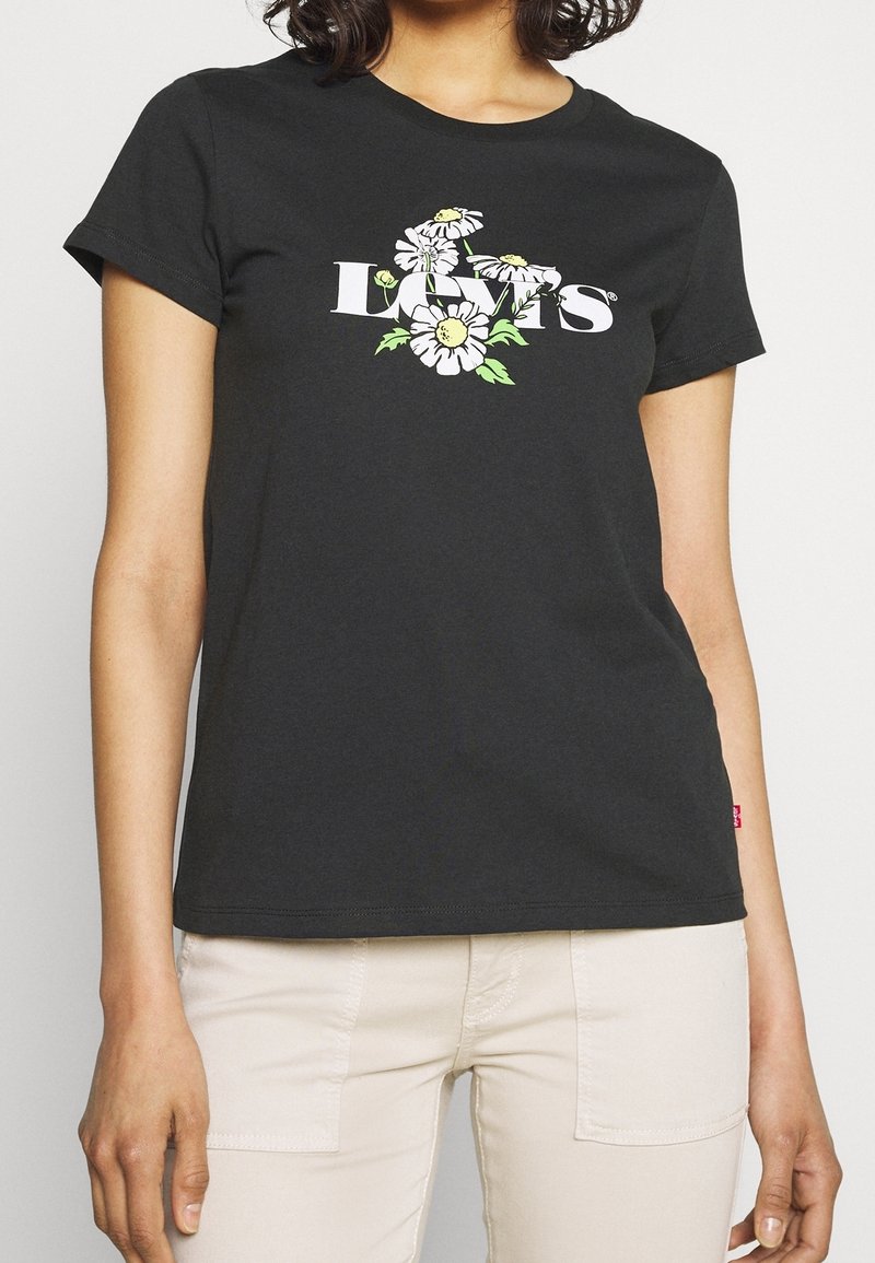 Femme portant un t-shirt Levi's noir avec un logo floral blanc et jaune, associé à un pantalon beige clair.