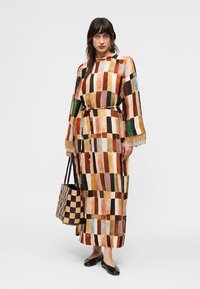 Γυναίκα που φοράει μακρύ φόρεμα με πολύχρωμο ορθογώνιο σχέδιο και φούντες στα μανίκια, κρατώντας μια τσάντα ώμου με μοτίβο καρό, στέκεται μπροστά σε λευκό φόντο.