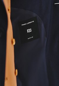 Ciemna tkanina odzieży z beżowym guzikiem. Posiada czarną metkę z napisem "PAKO LORENTE", "PJ" i "BLACK LINE" w białym tekście.