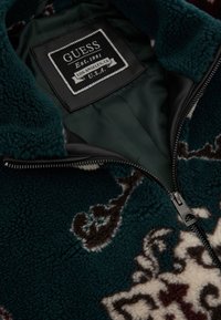 Žalias fliso džemperis su floraliniu raštu rudu ir dramblio kaulo spalvos. Jame yra odinė etiketė ir užtrauktukas, o vidinė pamušalo dalis yra lygi.