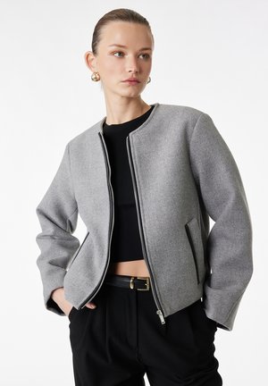 Donna che indossa una giacca grigia con zip sopra un top corto nero e pantaloni neri, con le mani nelle tasche, guarda di lato.