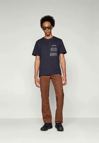 Navy blue t-shirt with graphic print on the chest; paired with brown pants featuring subtle stitching; model wears black sunglasses and shoes.