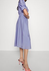 Femme debout, bras croisés, portant une robe midi violet clair et des sandales à talons roses sans brides, sur un fond uni.