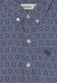 Camisa de manga corta con botones en color púrpura oscuro con un patrón floral geométrico, que presenta un pequeño logo bordado de alce y botones blancos.