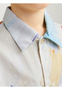 Lähikuva henkilöstä, jolla on päällään vaalean värinen paita, jossa on monivärinen akvarellikuvio ja napitettu kaulus.