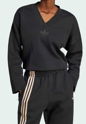 Person trägt schwarzen V-Ausschnitt-Sweatshirt mit Adidas-Logo und passende schwarze Hose mit beigen dreifachen Streifen an den Seiten, die Hände in den Taschen.
