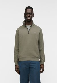 Φούτερ με φερμουάρ σε ελιά πράσινο χρώμα, με ψηλό γιακά, ριμπ μανσέτες και τελείωμα· διαθέτει ένα μικρό λογότυπο στο στήθος.