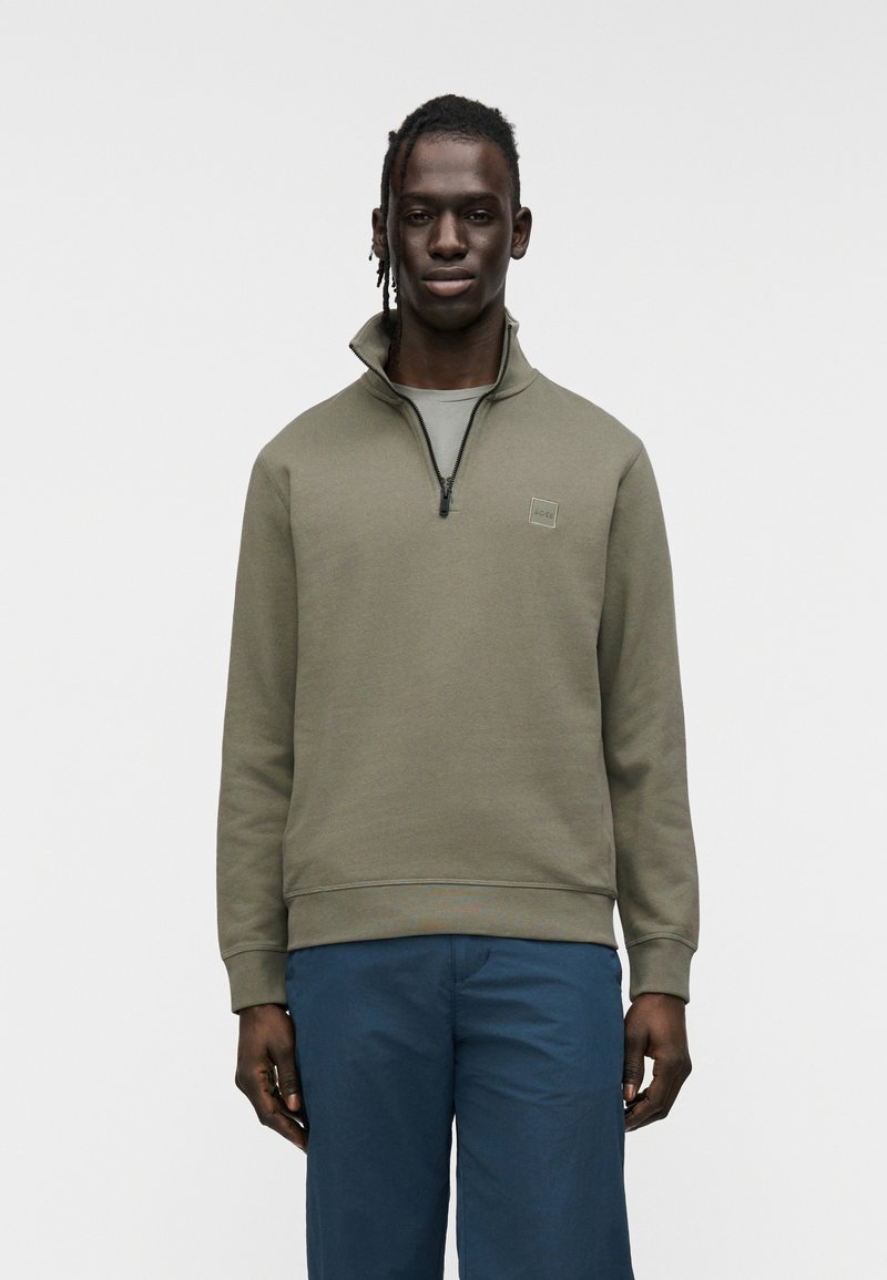 Φούτερ με φερμουάρ σε ελιά πράσινο χρώμα, με ψηλό γιακά, ριμπ μανσέτες και τελείωμα· διαθέτει ένα μικρό λογότυπο στο στήθος.