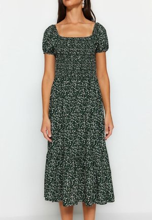 Femme portant une robe midi vert foncé avec un motif floral blanc, un décolleté carré et des manches courtes bouffantes.