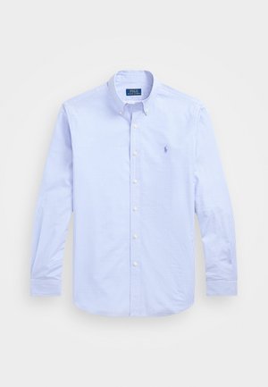 Šviesiai mėlyna marškininė su mygtukais, su languota raštą, ilgomis rankovėmis ir maža siuvinėta emblema kairėje krūtinės pusėje. Pagaminta iš medvilnės.