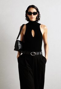 Top negro sin mangas con cuello alto que presenta un corte en forma de lágrima, combinado con pantalones negros de tiro alto y un pequeño bolso negro.