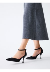 Zapatos de salón de raso negro con tacones altos, punta puntiaguda, pulsera en el tobillo con hebilla dorada y delgados tacones de aguja.