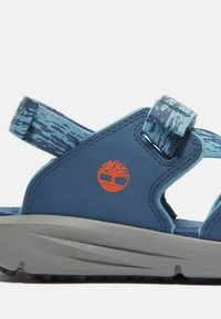 Siniset sandaalit, joissa on teksturoitu sininen hihna, harmaa pohja ja oranssi logo. Varustettu säädettävillä hihnoilla turvallista istuvuutta varten ja avoimella muotoilulla.
