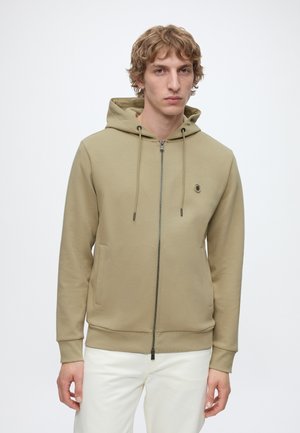 Blond man wearing a beige zip-up hoodie with drawstrings and white pants, standing against a plain white background.