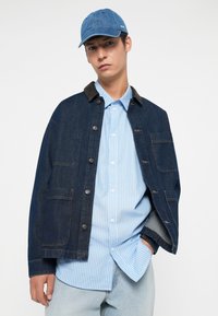 Denim jacket with dark blue color, brown collar, and front pockets, layered over a light blue striped shirt. Blue cap completes the look.