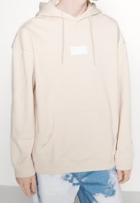 Beige hupparipaita, jossa on nyörit, kengurutasku ja pieni suorakulmainen etiketti. Pehmeä pinta ja rento istuvuus.