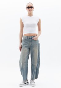 Bílý ribovaná crop top s krátkými rukávy, kombinovaný s volnými světle modrými džíny s vybledlou strukturou a viditelnými švy. Bílé tenisky.