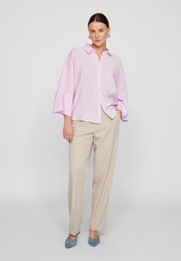 Lys rosa skjorte med brede ermer, kombinert med beige bukser og blå mules. Stoffet ser jevnt ut med en avslappet passform.