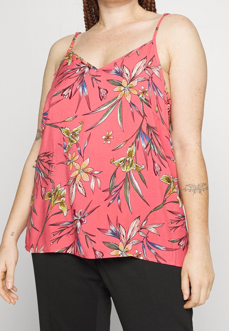 Femme portant un haut sans manches rose à fleurs avec des bretelles fines et un pantalon noir, montrant des tatouages sur les bras et les cheveux tressés.