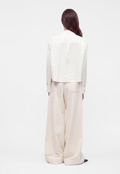 Femme aux longs cheveux portant une chemise blanche à manches longues et un pantalon large beige, debout de dos devant un fond uni.