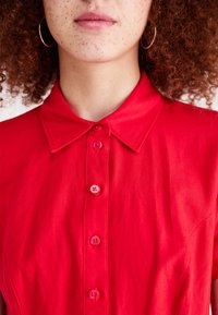 Κόκκινο πουκάμισο με κολάρο και κουμπιά στο μπροστινό μέρος, διαθέτοντας πέντε ταιριαστά κουμπιά. Απαλή υφή, κοντά μανίκια και ορατές λεπτομέρειες ραφής.
