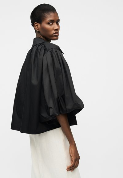 Femme portant un chemisier noir ample à manches bouffantes associé à une jupe de couleur claire, debout devant un fond uni.