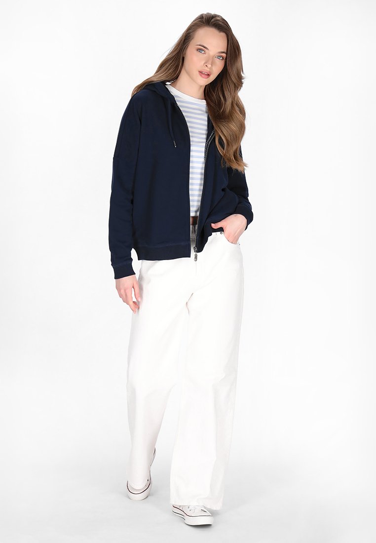 Marineblauer Zip-Hoodie mit einer Fronttasche, kombiniert mit einem hell gestreiften Hemd und weiten weißen Hosen. Das Outfit wird mit weißen Sneakern abgerundet.