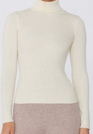 Pull à col roulé côtelé de couleur crème avec manches longues, porté avec un pantalon doux couleur taupe.
