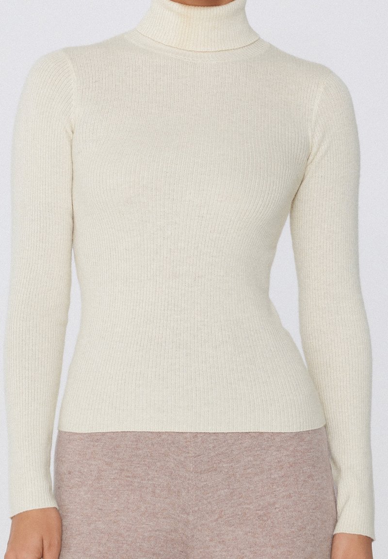 Pull à col roulé côtelé de couleur crème avec manches longues, porté avec un pantalon doux couleur taupe.