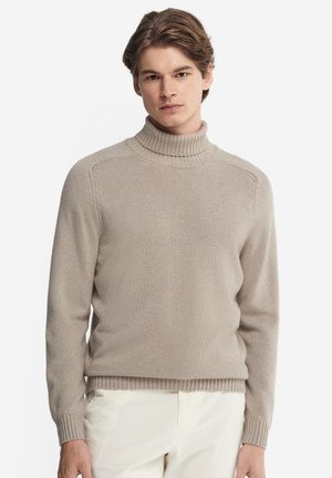 Młody mężczyzna w beżowym golfie i białych spodniach, stojący na jednolitym białym tle.