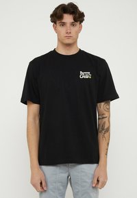 Joven con cabello corto y castaño lleva una camiseta negra con el texto "Barrow The Love For Crew" y pantalones gris claro, mostrando tatuajes en el brazo derecho.