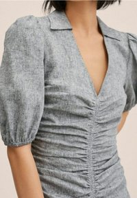Femme portant une robe grise à fronces avec des manches bouffantes et un col en V profond.