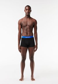 Boxer-briefs noirs avec une ceinture bleue portant le texte "LACOSTE". Le tissu est lisse et le design est ajusté avec un devant en contour.