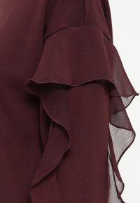 Μπορντό μπλούζα με στρώσεις ριγέ μανικιών και ημιδιάφανη και υφασμάτινη επιφάνεια, σε συνδυασμό με μια λεία βάση.