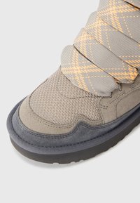Világosszürke sneaker, textúrált felsőrésszel, velúr részletekkel és narancssárga kockás pánttal. Lekerekített orr és tartós fekete gumitalp.