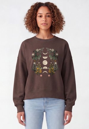 Junge Frau mit lockigem Haar trägt einen braunen Pullover mit Blumen- und Mondphasen-Design, kombiniert mit hellblauen Jeans, vor weißem Hintergrund.