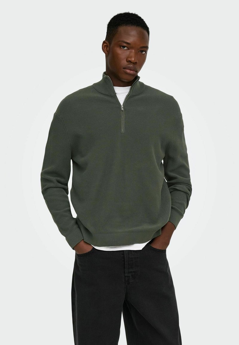 Jeune homme portant un pull zippé vert foncé par-dessus une chemise blanche, les mains dans les poches, debout devant un fond clair uni.